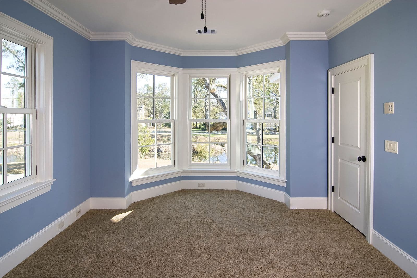 Bright blue bedroom with bay window overlooking landscaped yard, carpeted floor, white trim, and cream-colored double doors on right wall