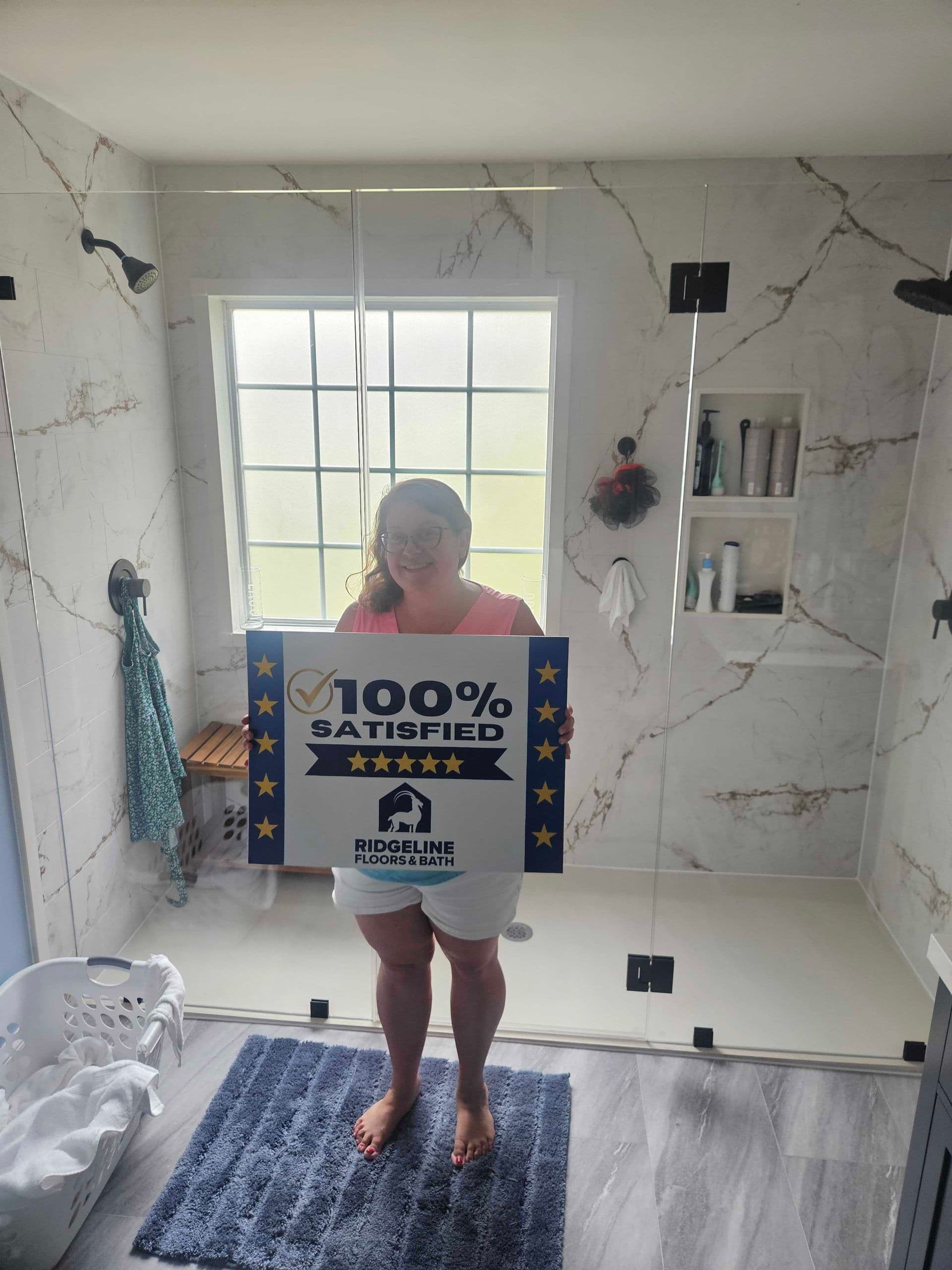 Satisfied customer holding a 100% satisfied five-star review sign for Ridgeline Floors & Bath in a newly renovated marble bathroom with glass shower enclosure