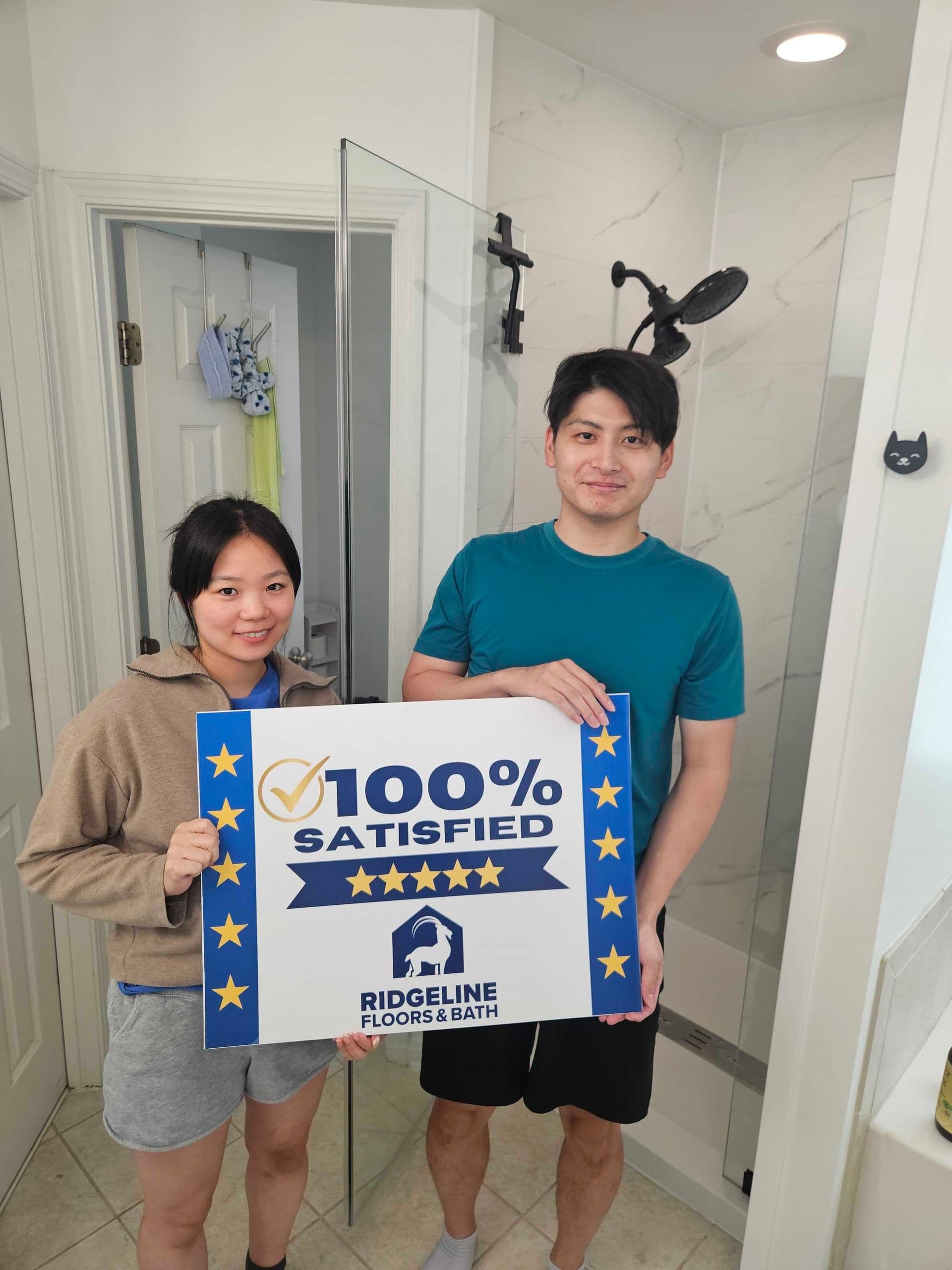 Two people holding a 100% Satisfied five-star rating sign for Ridgeline Floors & Bath in a modern bathroom with marble walls and glass shower enclosure
