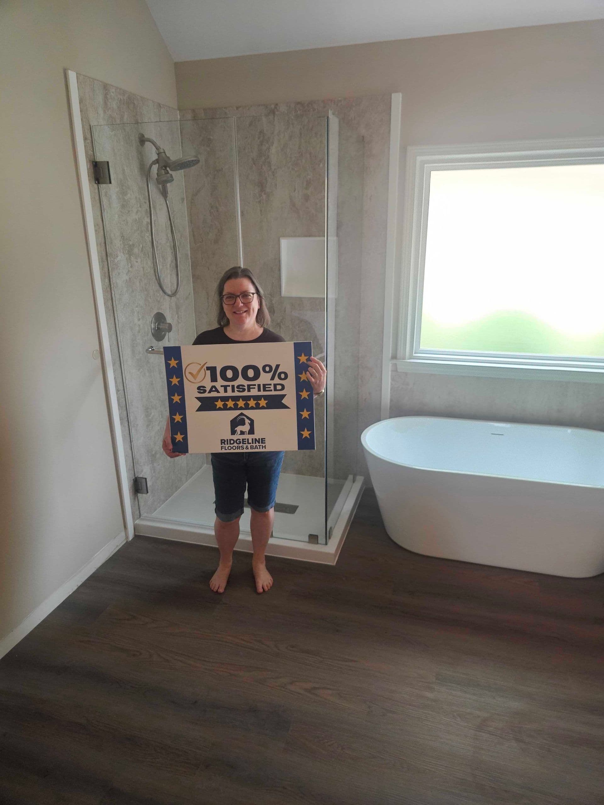 Woman holding a 100% satisfied customer review sign in a bathroom with a glass shower enclosure and freestanding tub