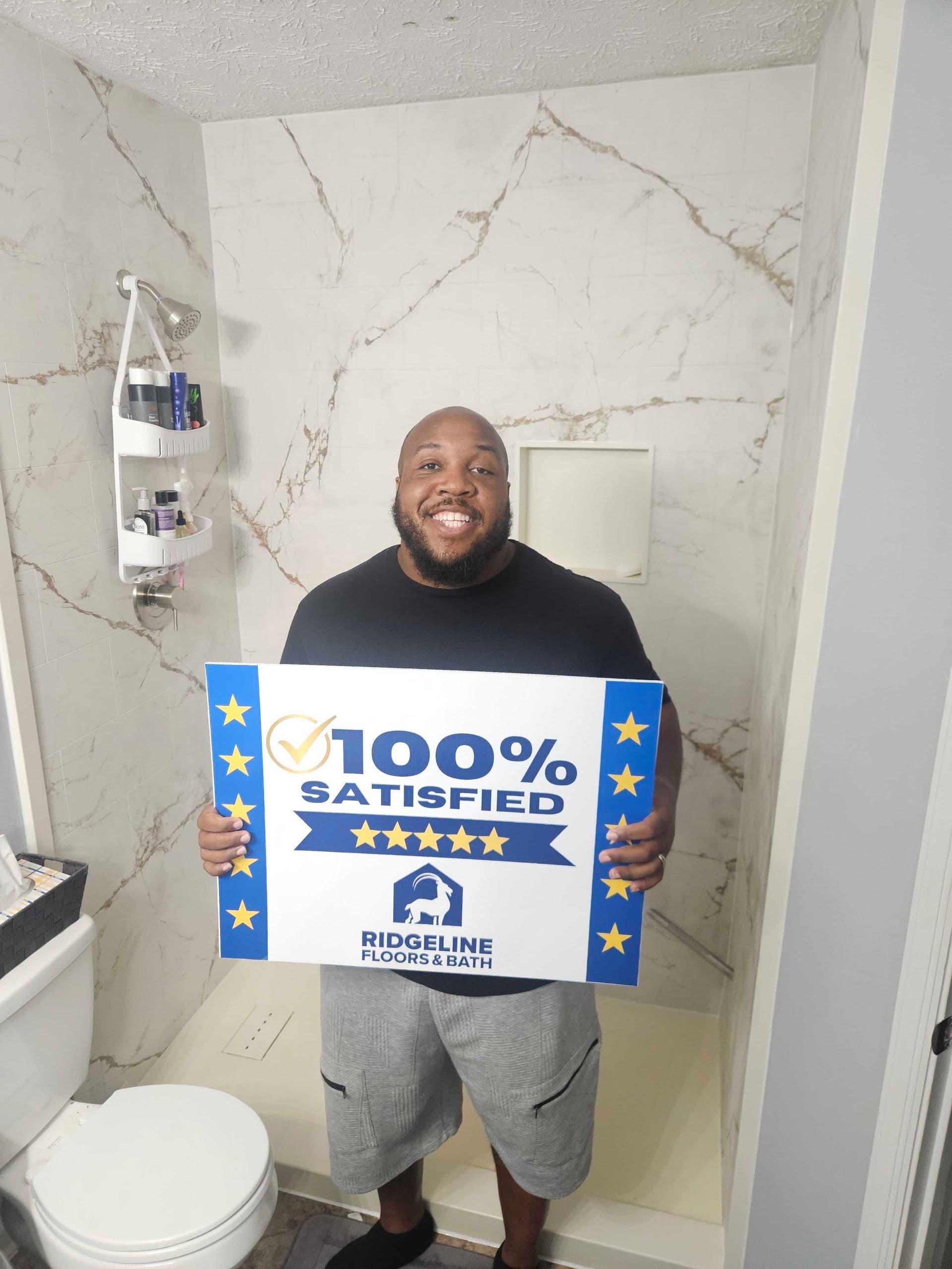 Man holding a 100% satisfied customer review sign for Ridgeline Floors & Bath in a bathroom with marble tile walls