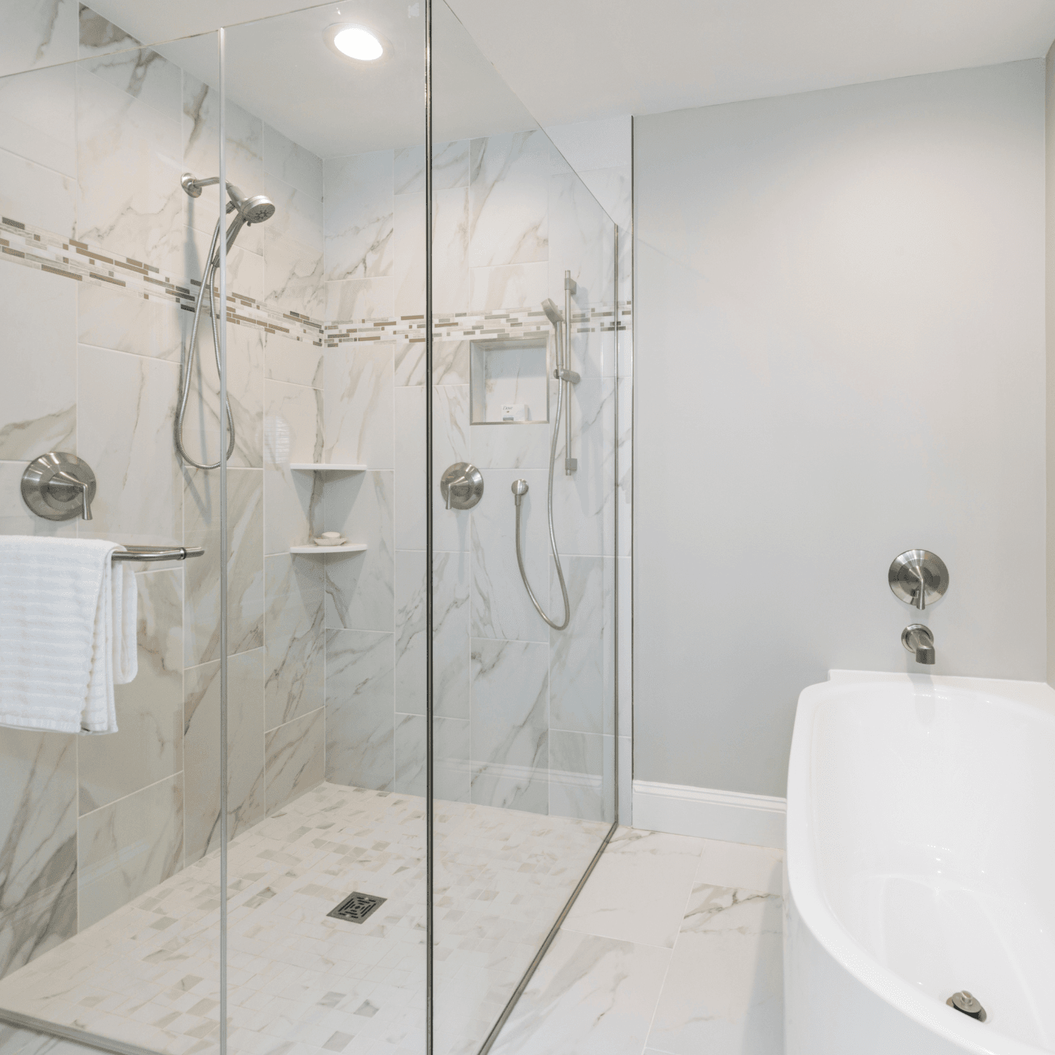 Modern bathroom with walk-in shower featuring marble tile walls, glass enclosure, dual showerheads, and built-in shelving, plus freestanding white soaking tub on right side