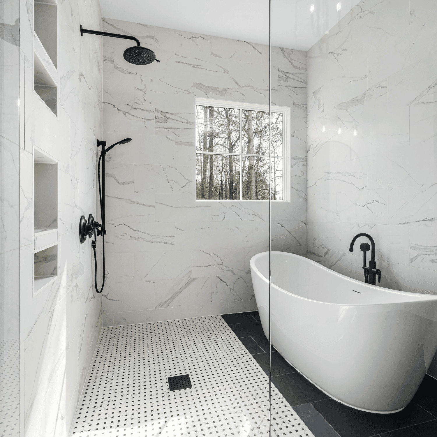 Modern luxury bathroom with marble-tile walls, freestanding white soaking tub, walk-in shower with glass partition, black fixtures, and window overlooking trees