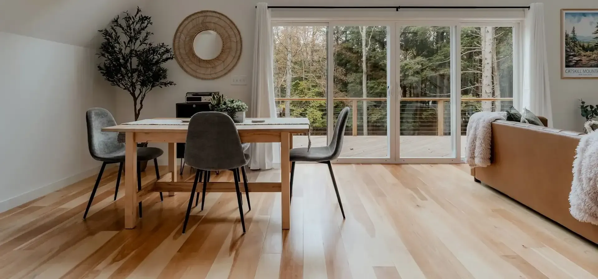 Modern open-concept living and dining space with wooden table, black chairs, tan sofa, large windows and glass doors overlooking forested landscape