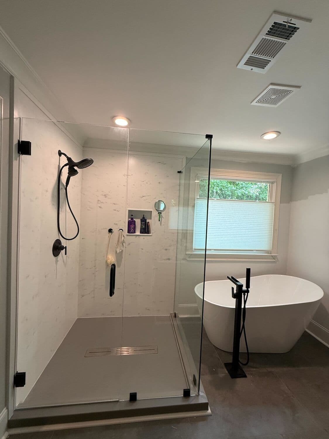 Modern bathroom with walk-in shower with marble tile walls and black fixtures on left, freestanding white soaking tub with black faucet on right, and window with blinds in background